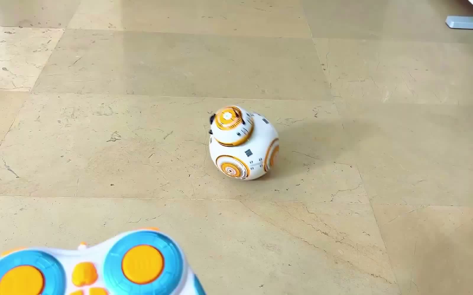 The droid toy driving across a tiled floor with the remote control held in the foreground