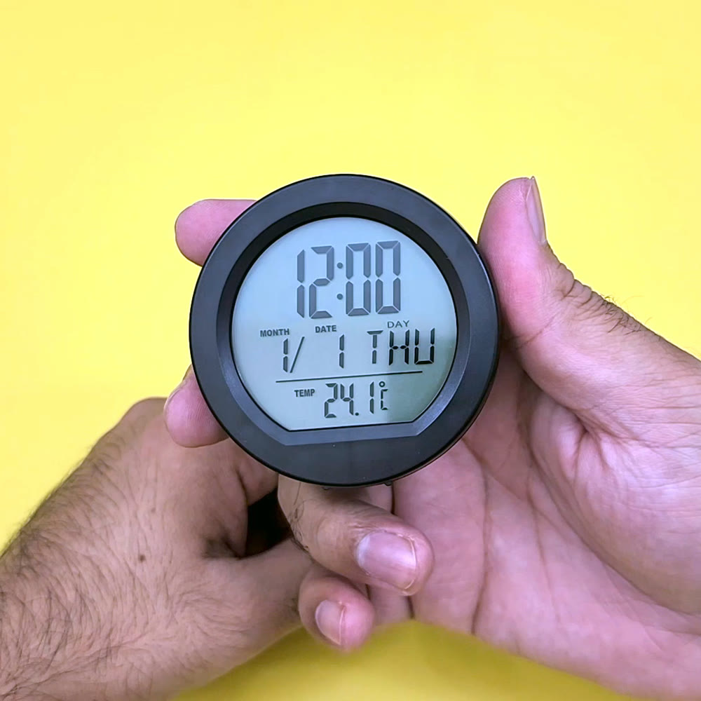 Round black solar car dashboard clock held in a hand, its LCD showing the time, the day and date and the temperature in Celsius