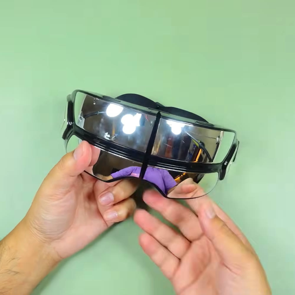 Oaks eye massager held front-on in two hands, the mirrored visor panels facing the camera and the strap hanging below