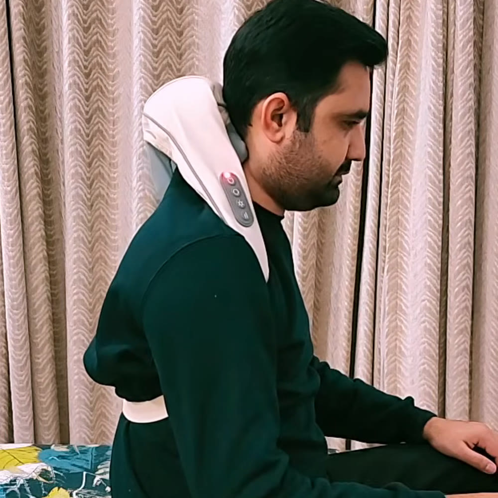 Man sitting on a bed wearing the neck and shoulder massager over his shoulders, the arm straps hanging down his chest