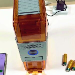 The actual Ostation X charger standing on a white desk beside the phone app and three loose AA batteries