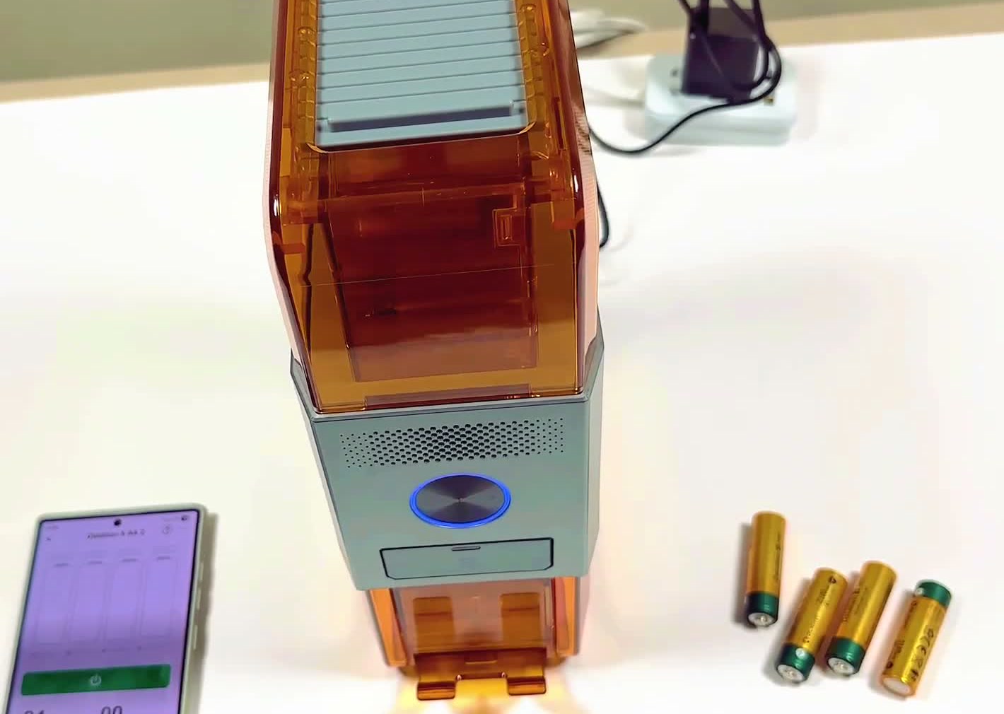 The actual Ostation X charger standing on a white desk beside the phone app and three loose AA batteries