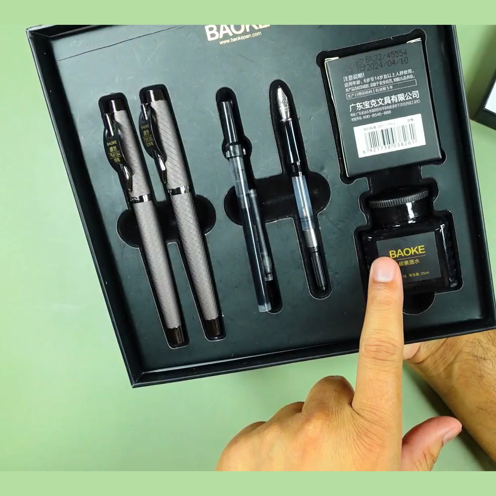 Open BAOKE fountain pen gift box on a desk, holding four metal pens in fitted foam beside a boxed bottle of ink