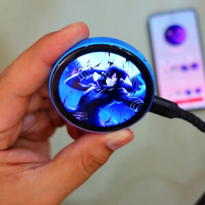 Blue digital anime pin held in one hand, its round HD touchscreen showing anime artwork, with the companion phone app open behind it