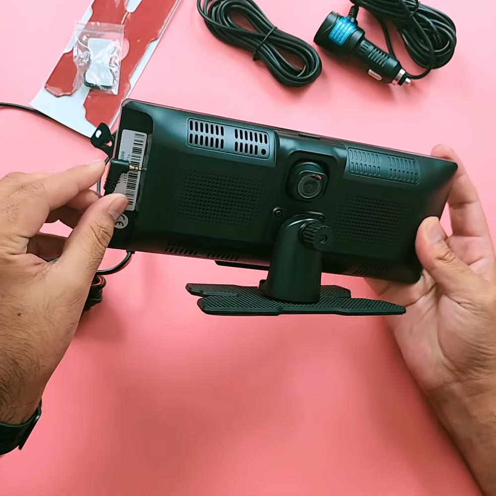 Back of the PODOFO A3073 held in two hands, showing the built-in front dash cam lens, the speaker grilles and the swivel stand