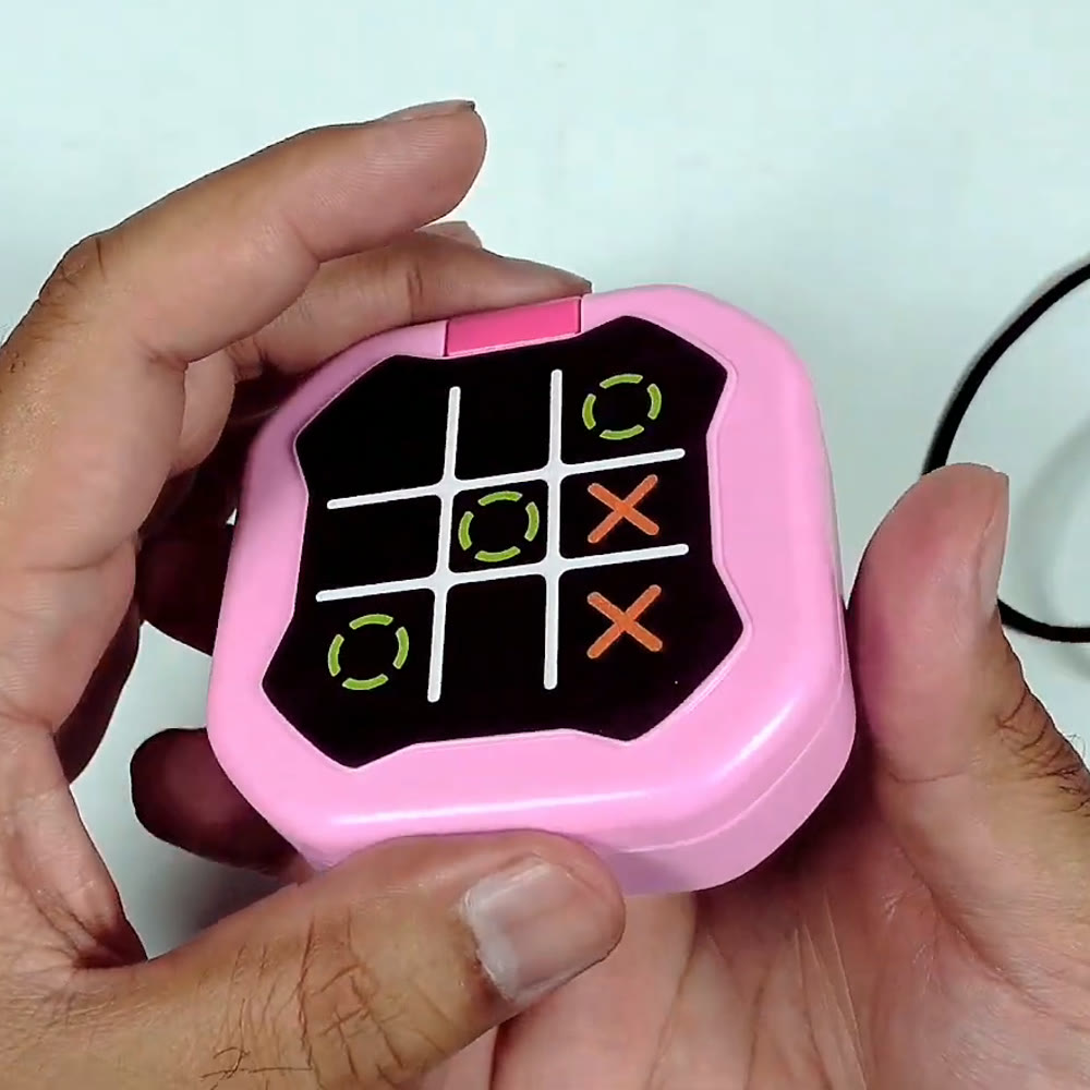 Pink handheld digital tic tac toe game held closed in hand, showing the X and O grid artwork on the lid and its wrist lanyard